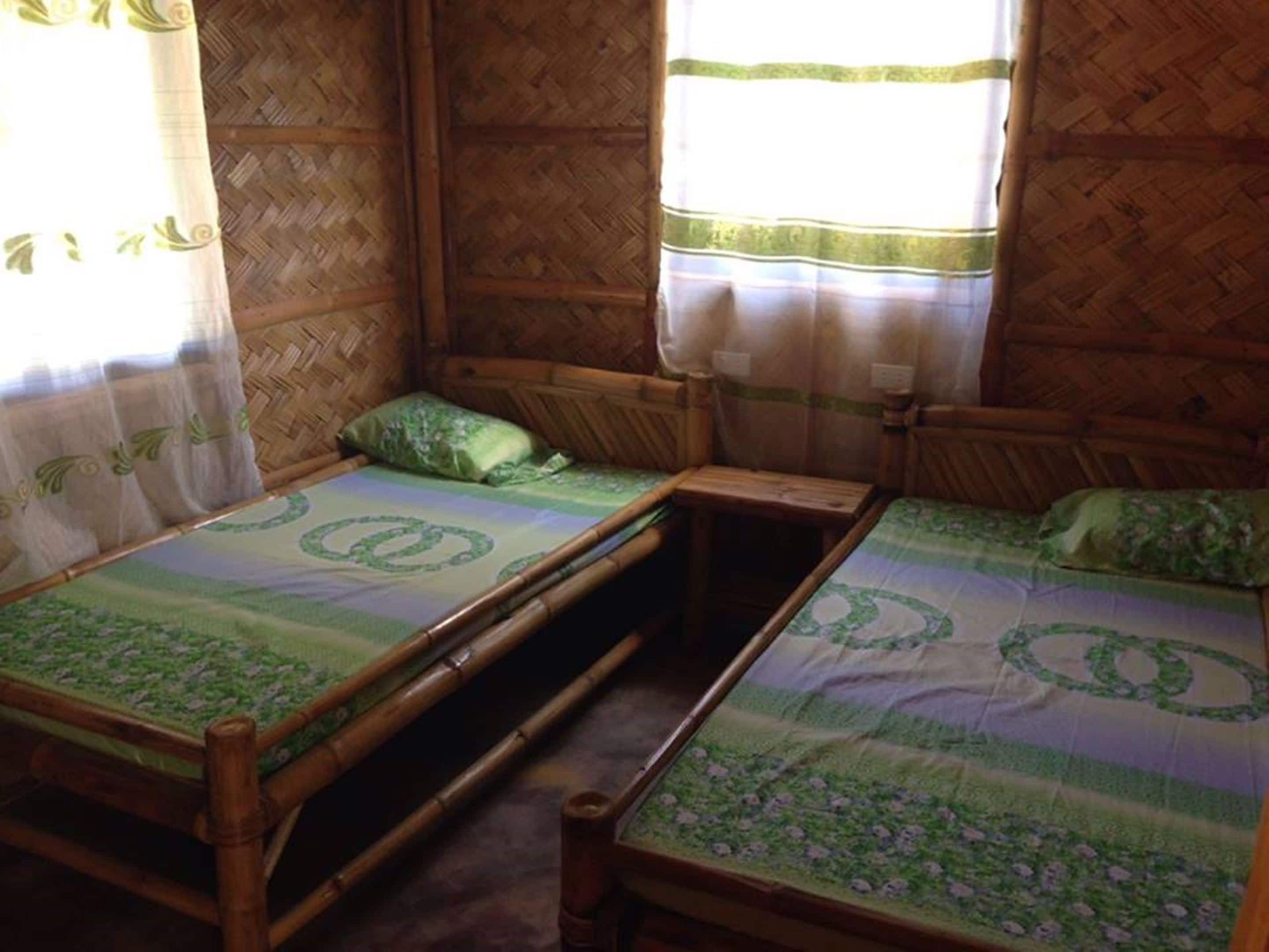 A room with two wooden beds, each with a green patterned bedspread and matching pillows. The room has a woven bamboo wall and a window with sheer curtains.