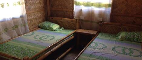 A room with two wooden beds, each with a green patterned bedspread and matching pillows. The room has a woven bamboo wall and a window with sheer curtains.