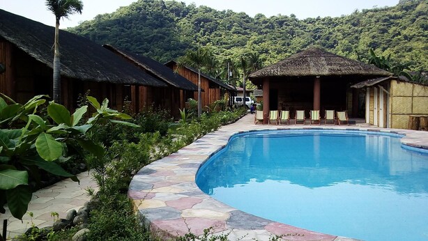 Zona junto a una piscina bordeada de piedra, edificios de madera con techos de paja y abundante vegetación al fondo.
