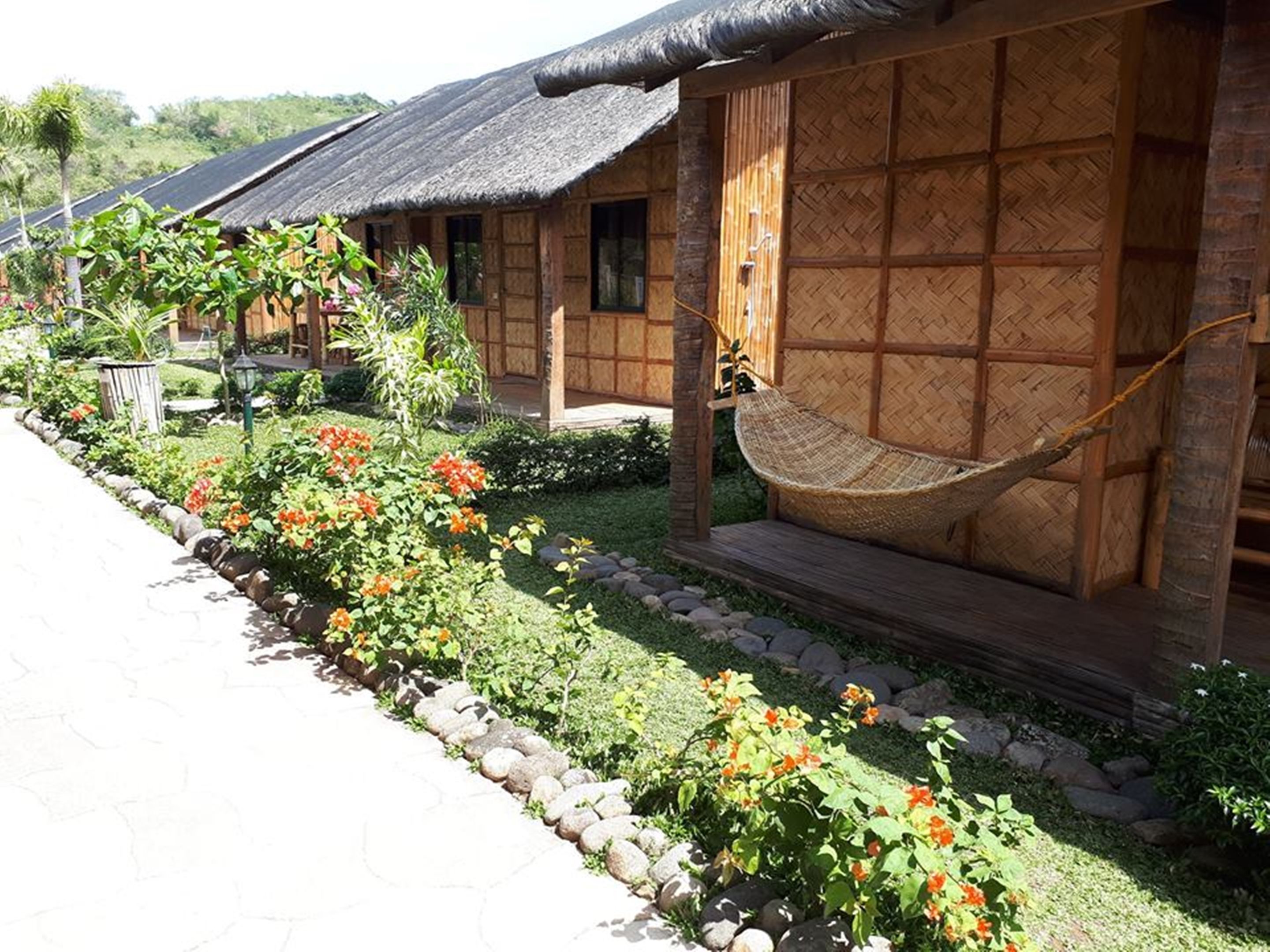 A row of traditional wooden houses with thatched roofs, a hammock, and a garden with orange flowers.