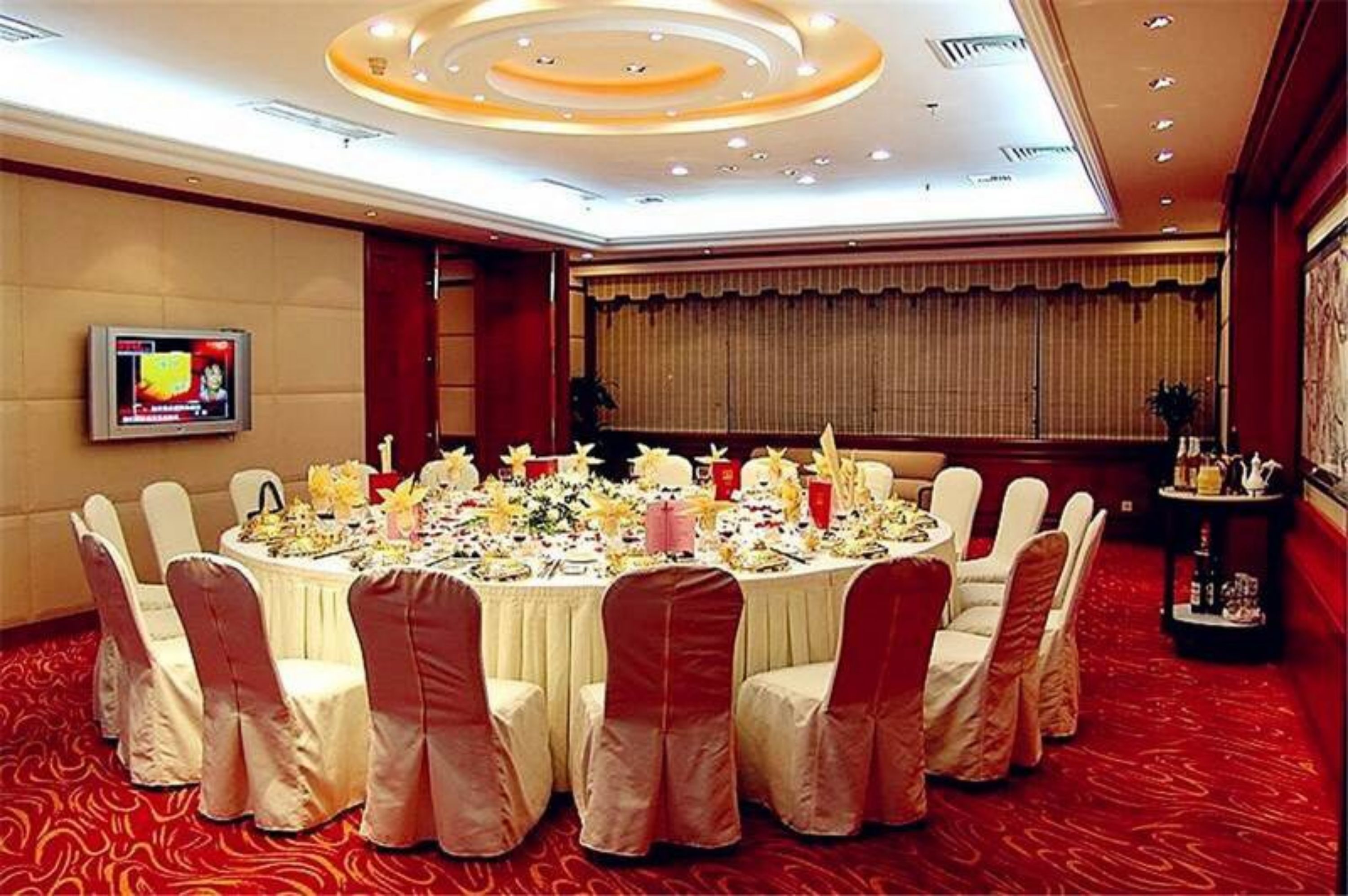A conference room with a round table set for a meeting, surrounded by chairs with red sashes, a television on the wall, and a red carpeted floor.
