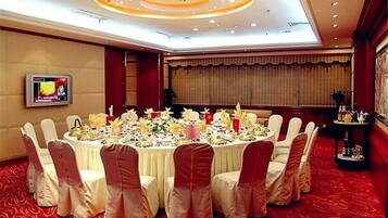 A conference room with a round table set for a meeting, surrounded by chairs with red sashes, a television on the wall, and a red carpeted floor.