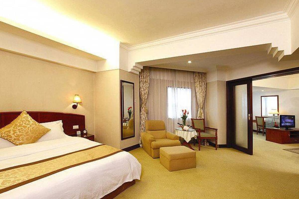 A hotel room with a large bed, a sitting area with a chair and ottoman, a desk with a lamp, and a television.