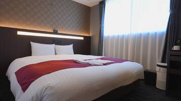 Habitación de hotel con una cama grande, una manta roja, almohadas blancas, cabecera con panel de luz, mesita de noche y un cubo de basura.