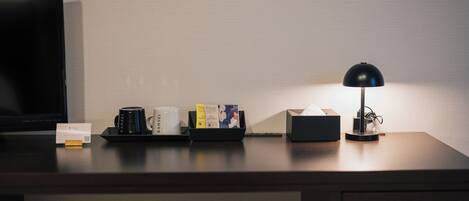 A hotel desk with a flat-screen TV, a lamp, a tray with a mug, a tissue box, and some brochures.