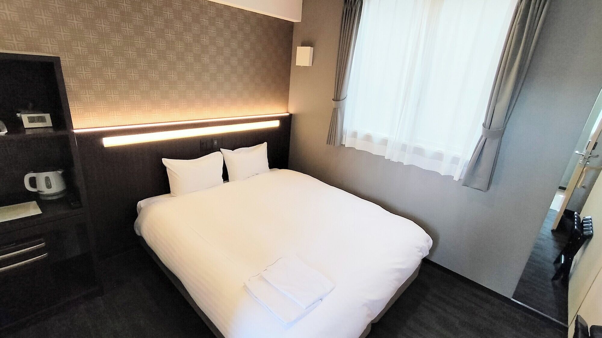 Een hotelkamer met een netjes opgemaakt bed, een nachtkastje met een waterkoker, een wandlamp en een raam met gordijnen.