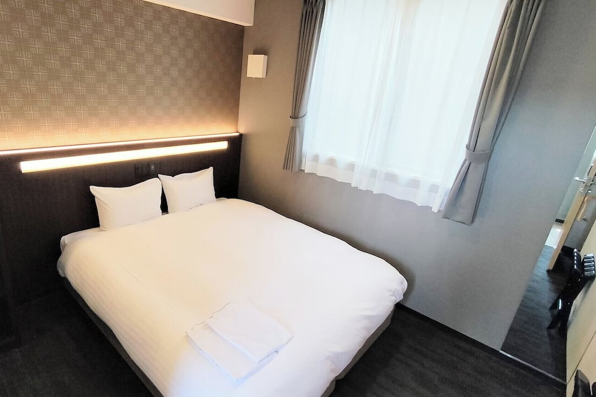 Ett hotellrum med en väl bäddad säng, ett sängbord med en vattenkokare, en väggmonterad lampa och ett fönster med gardiner.