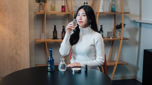 A person seated at a round table with a bottle, a glass, and a container. Behind them is a wooden shelf with various bottles. - KANSEI Kyoto Hachijo (Kyoto)