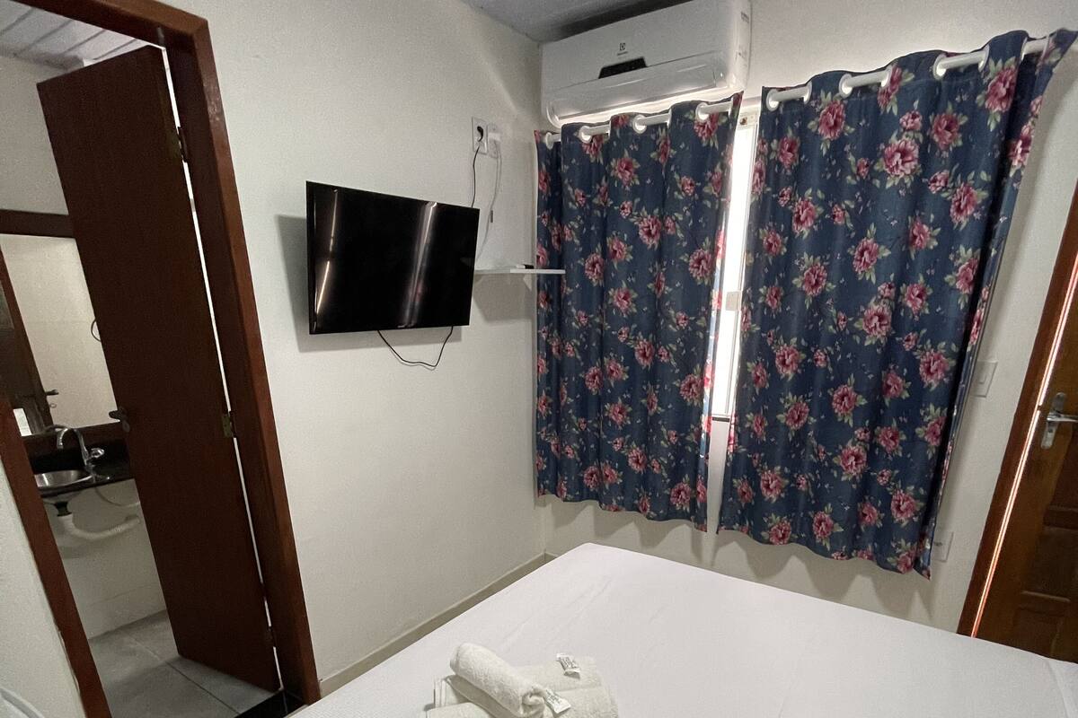 Ein Hotelzimmer mit Bett, Wandfernseher, sichtbarem Badezimmer durch eine geöffnete Tür und einem Fenster mit blumigem Vorhang.