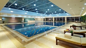 An indoor swimming pool with a high ceiling, lounge area with sunbeds, and a view of the pool.