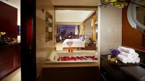 A modern hotel room with a large bed, a bathtub with rose petals, and a view of the city at night.