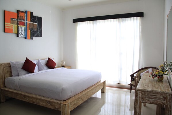 A bedroom with a wooden bed, white bedding, a nightstand, a desk with a chair, and a window with sheer curtains. - Ketut Villa Sanur (Denpasar)