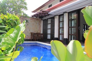 A pool area with a stone wall, wooden deck, and a table with chairs. - Ketut Villa Sanur (Denpasar)