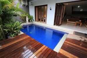 A swimming pool with a wooden deck, surrounded by lush greenery and a sliding glass door leading to a living area. - Nyoman Villa Sanur (Denpasar)