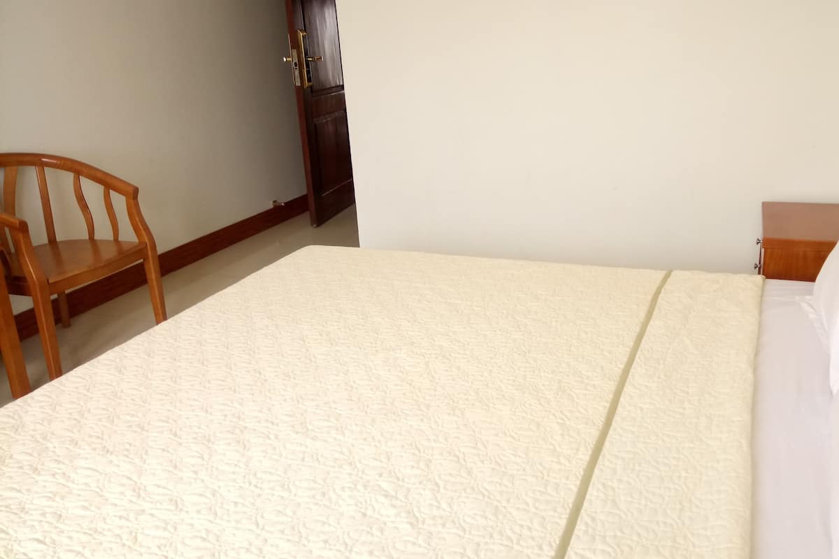 Una cama bien hecha con cubierta blanca y almohadas, una silla de madera y una mesita de noche con lámpara.