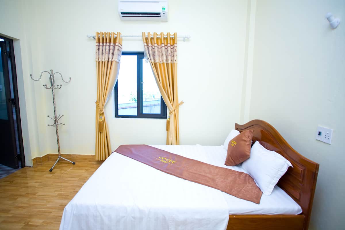 Una habitación de hotel con piso de madera, una cama con ropa de cama blanca y marrón, una lámpara de pie y una ventana con cortinas.