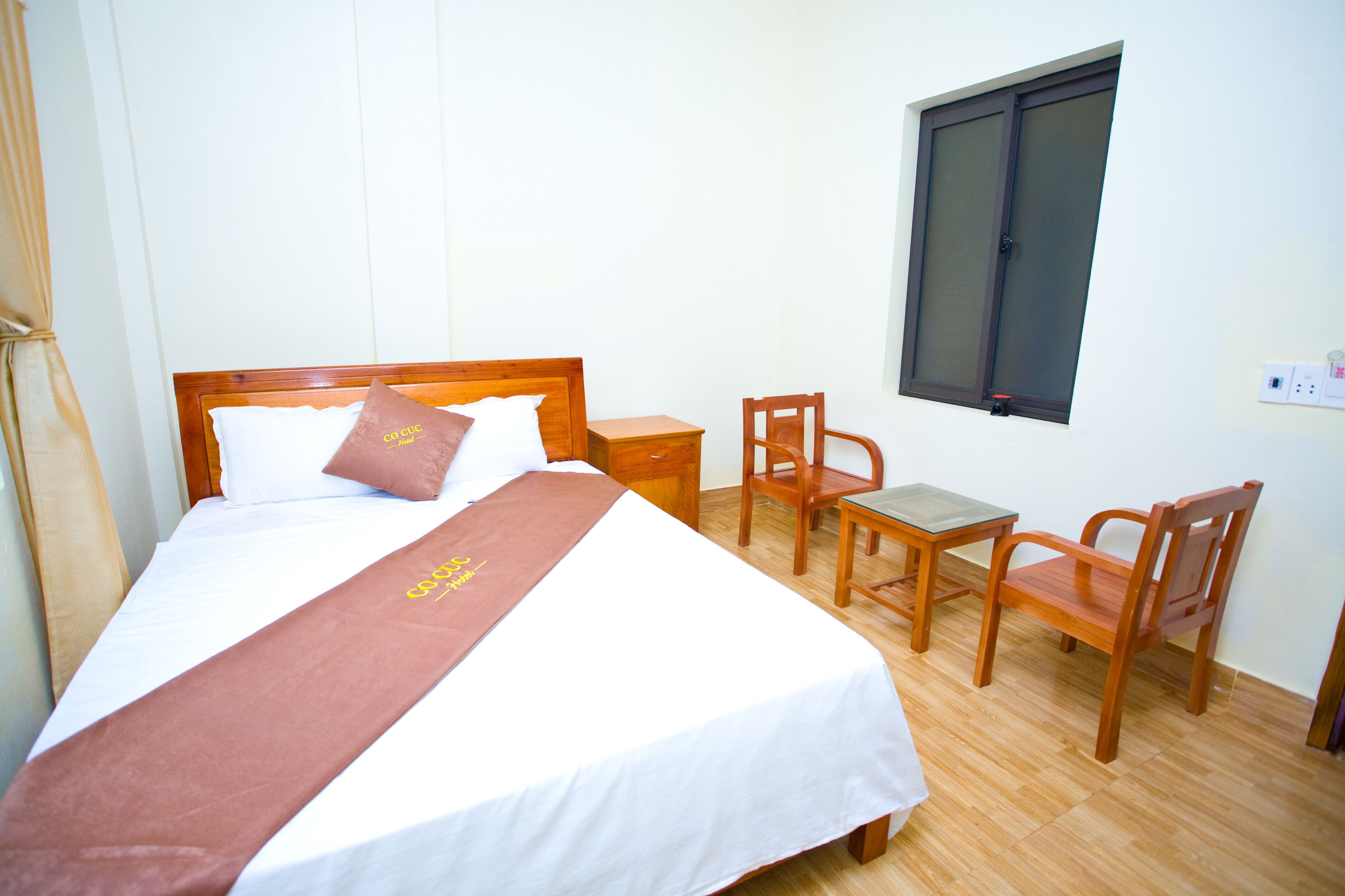 A neatly made bed with a brown bedspread, a wooden nightstand, two wooden chairs, and a window with blinds.