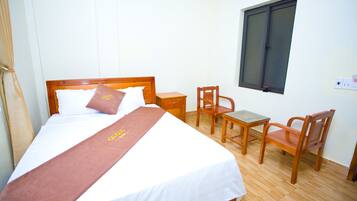 A neatly made bed with a brown bedspread, a wooden nightstand, two wooden chairs, and a window with blinds.