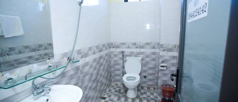 A bathroom with a white sink, toilet, and shower. There is a glass shelf above the sink and a small window above the toilet.
