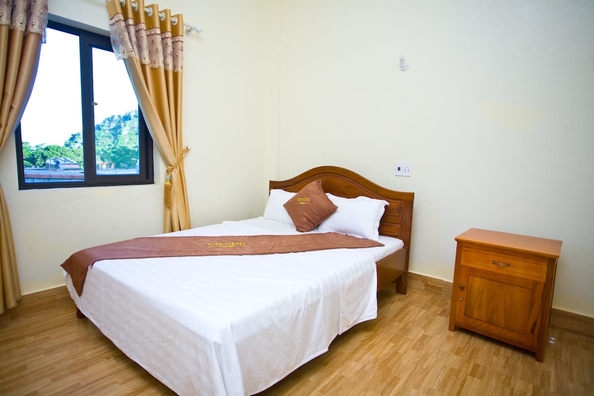Una cama bien hecha con un plaid marrón, una mesita de noche de madera y una ventana con cortinas.