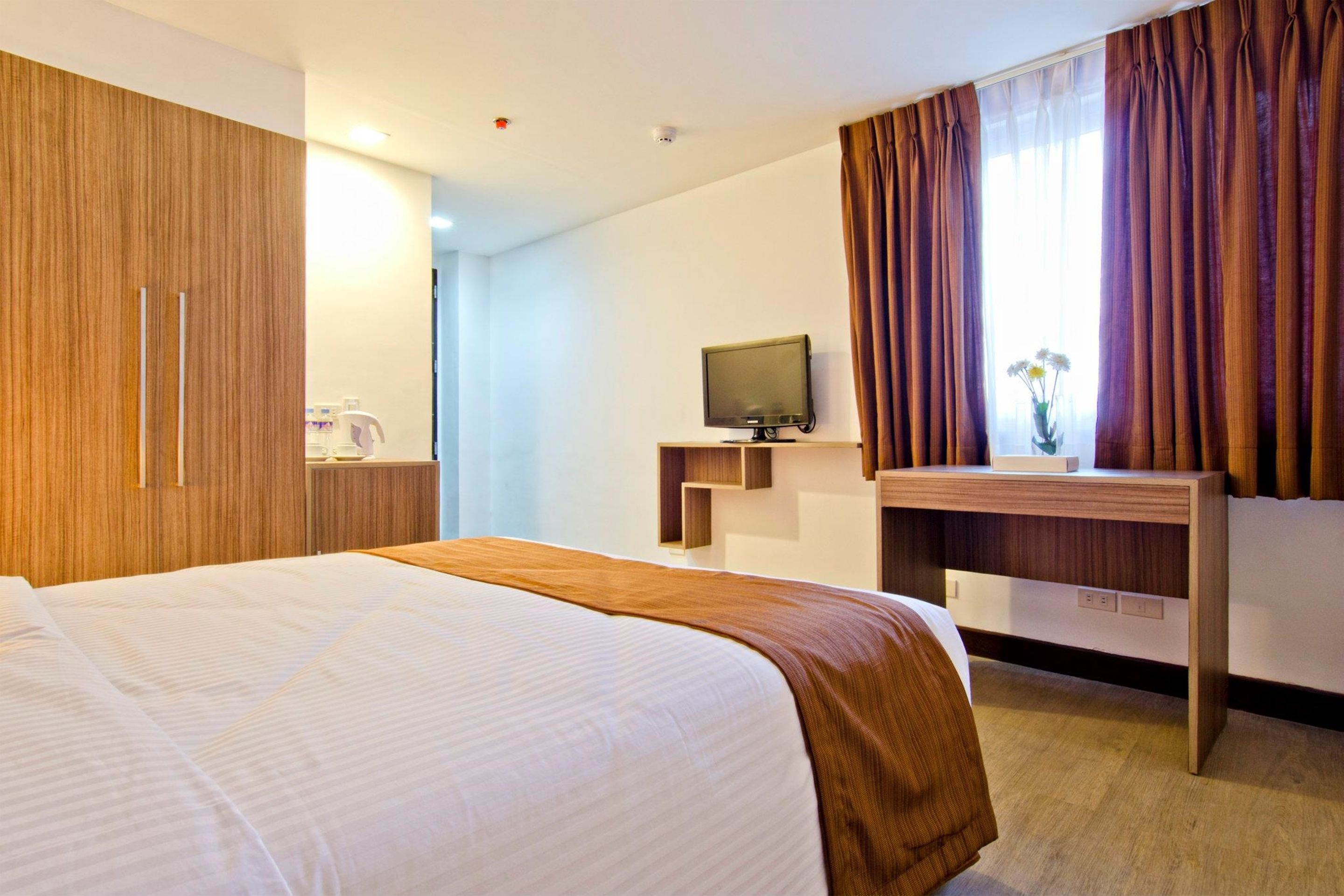 Habitación de hotel con una cama grande, un televisor sobre un soporte de madera, un escritorio con una computadora y una ventana con cortinas.
