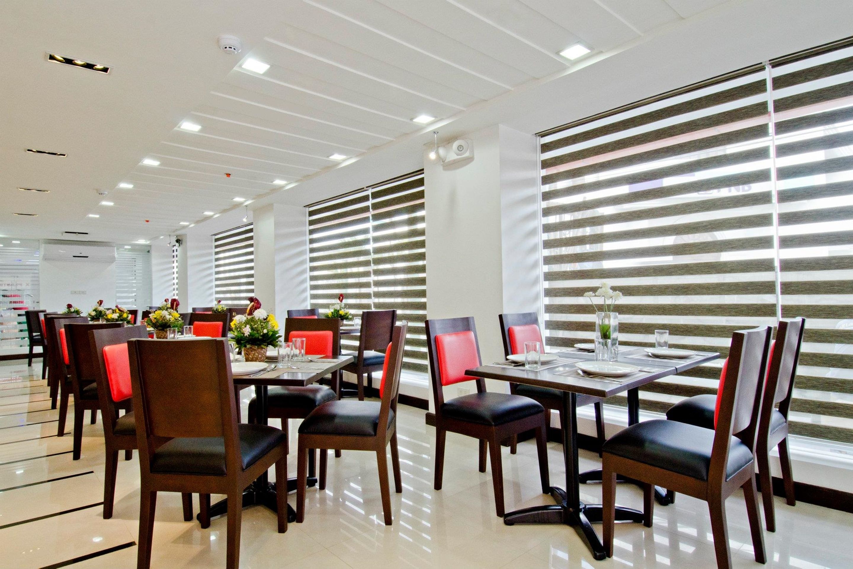Un comedor moderno con varias mesas y sillas, ventanas grandes con persianas e iluminación en el techo.