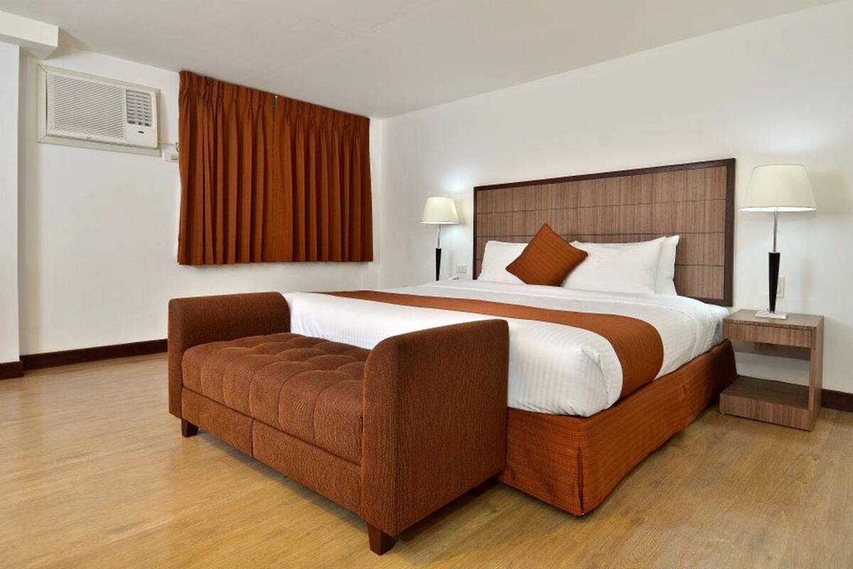 A hotel room with a large bed, a brown upholstered bench, two bedside tables with lamps, and a window with brown curtains.