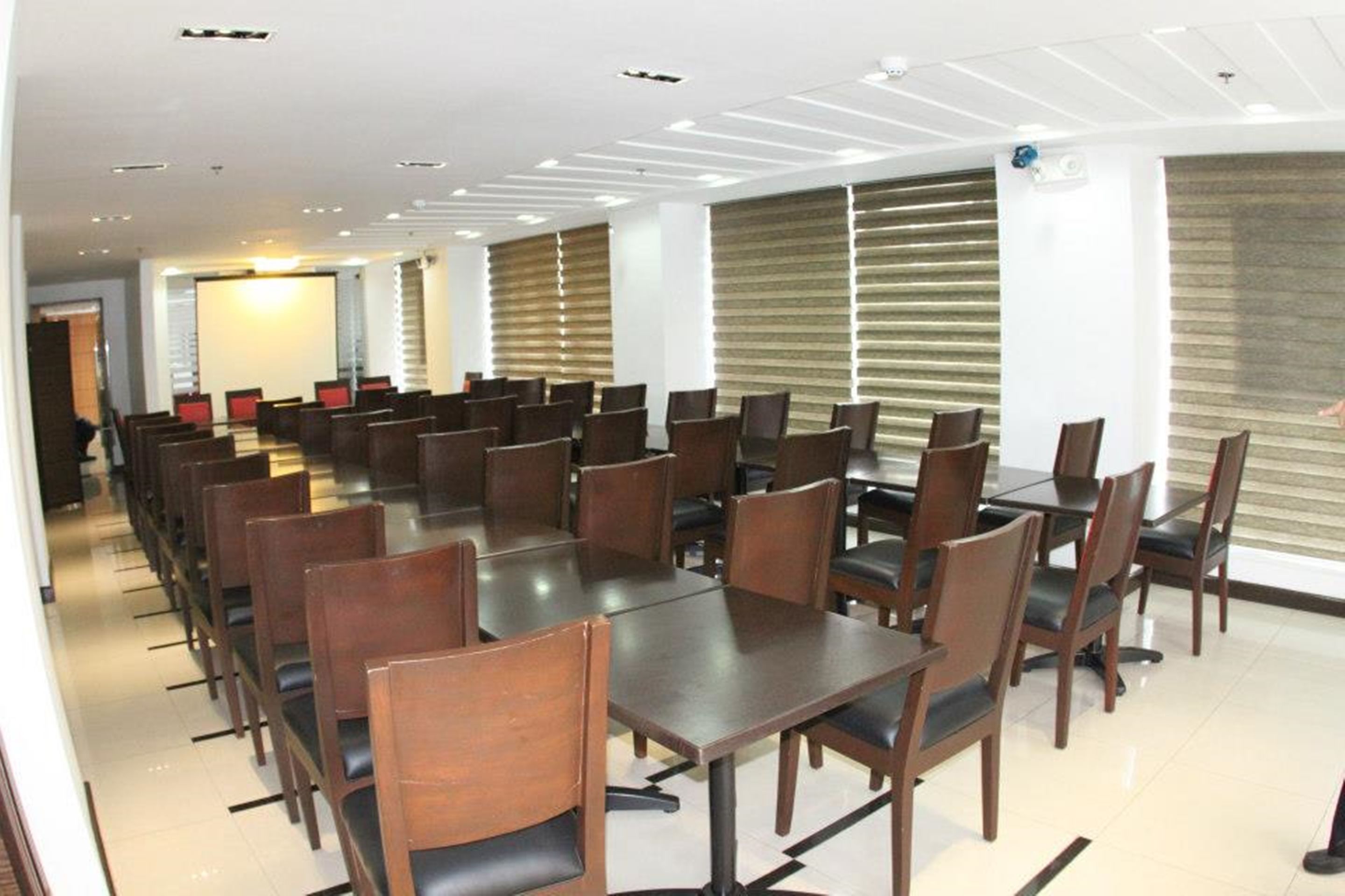 Sala de conferências com várias mesas e cadeiras, amplas janelas com persianas e design de teto moderno.