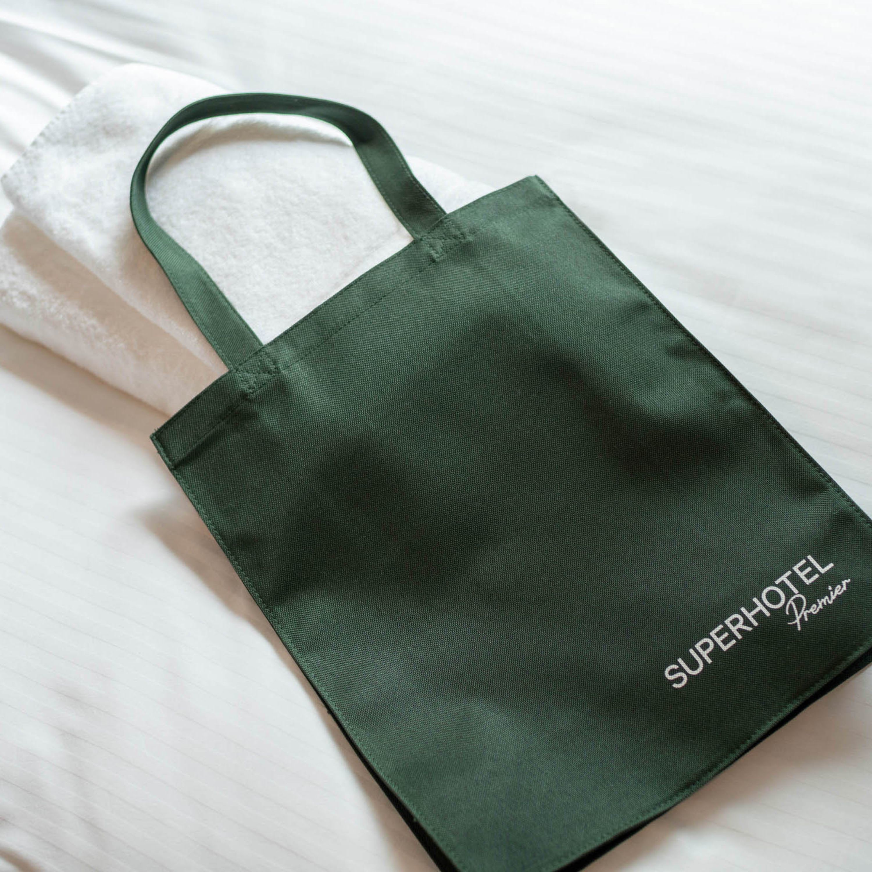 A green tote bag with the text "Superhotel Premier" on it, placed on a white bed.