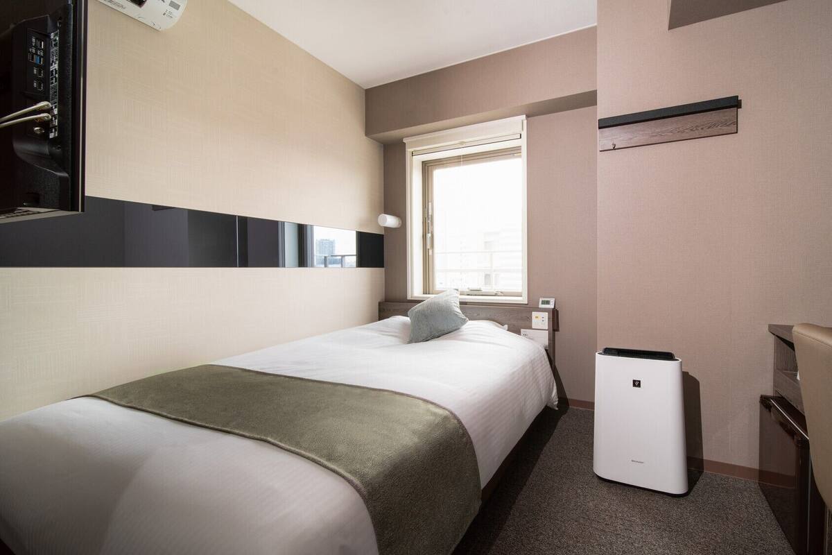 Una habitación de hotel con cama, escritorio, silla, una mesita, ventana y un dispositivo montado en la pared.