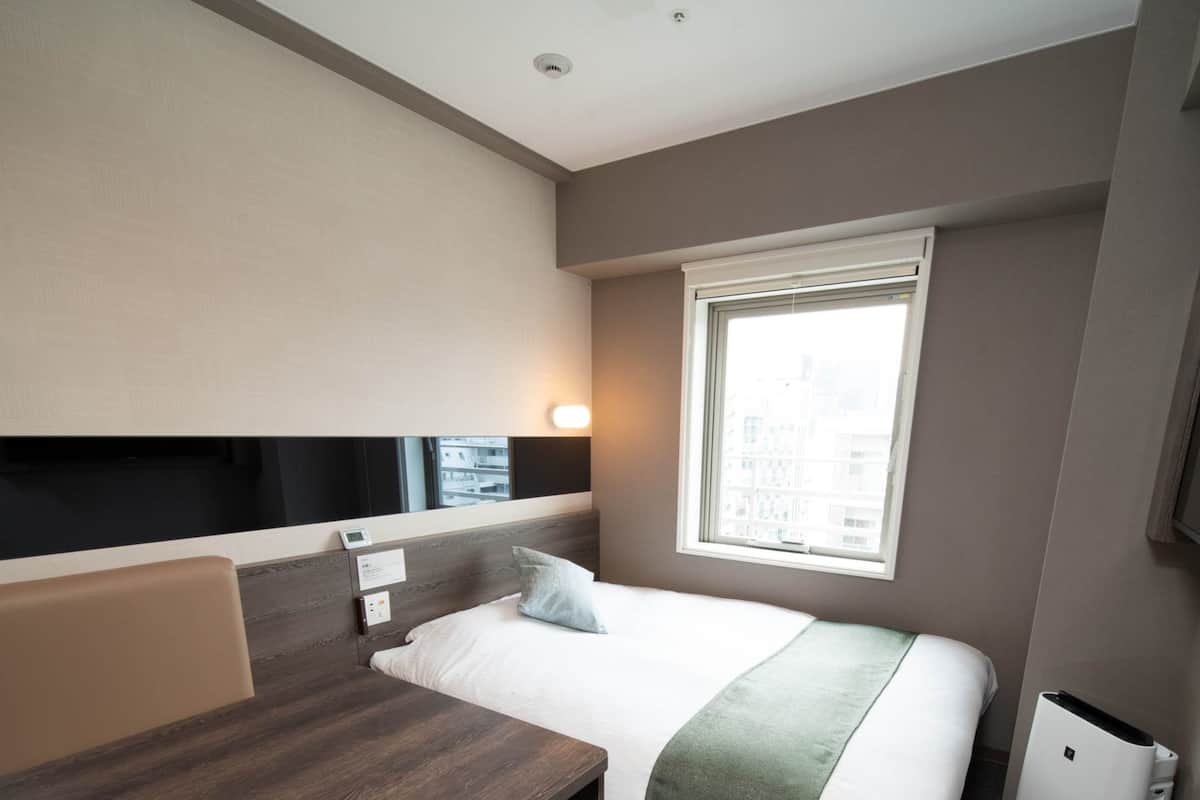 Habitación de hotel con cama, escritorio, silla, cubo de basura y ventana con vista a edificios.