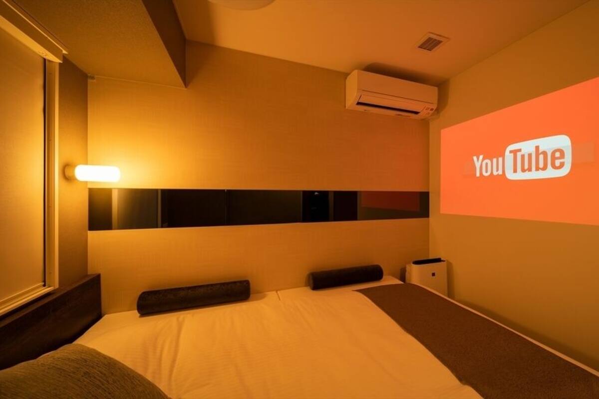 Habitación de hotel con cama, aire acondicionado de pared, mesita de noche y proyección del logotipo de YouTube en la pared.
