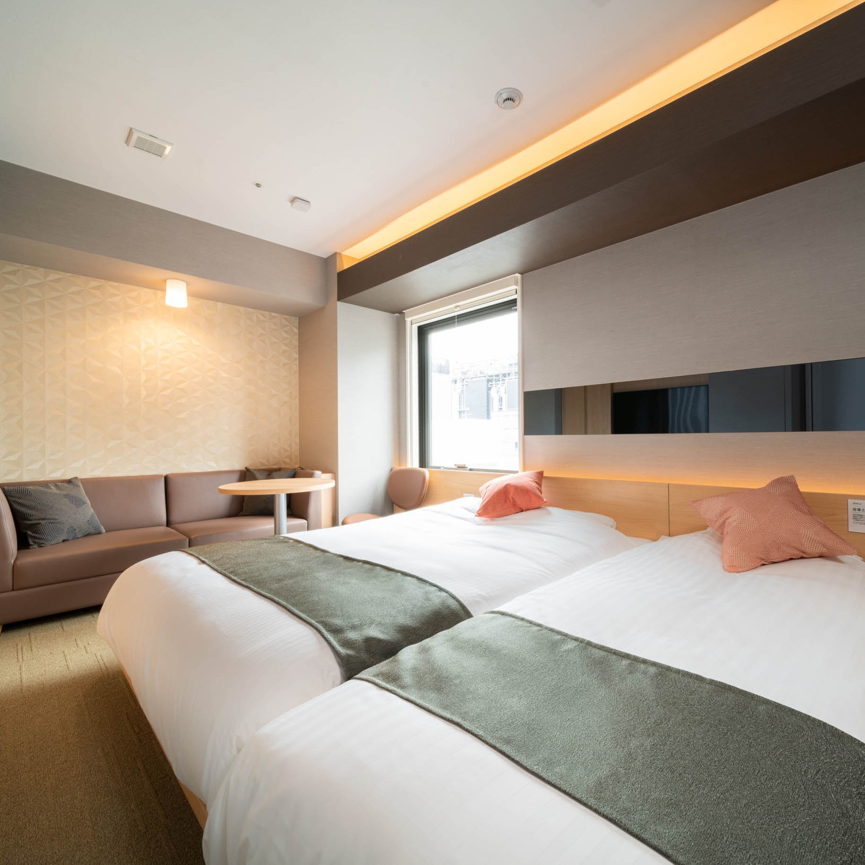 A modern hotel room with two beds, a sofa, a small table, and a window with a city view.