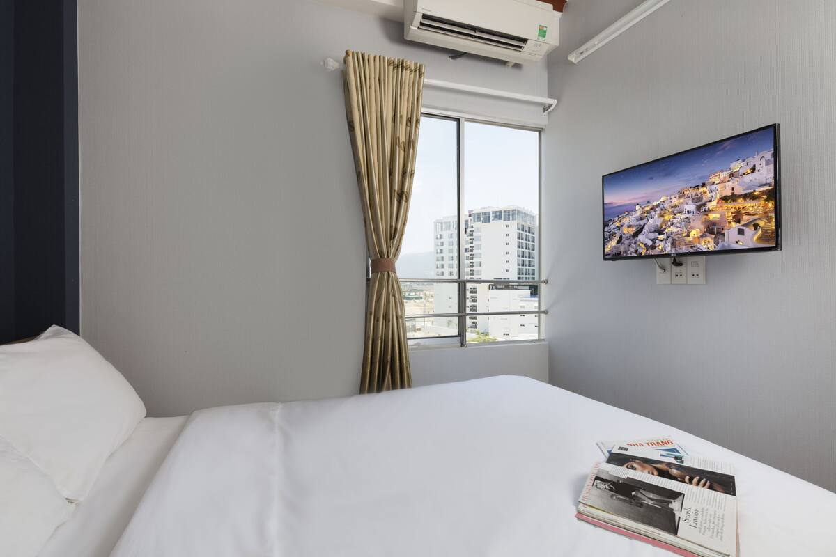Une chambre d’hôtel avec un lit, une fenêtre avec des rideaux, un climatiseur, un téléviseur fixé au mur et une vue sur le paysage urbain.