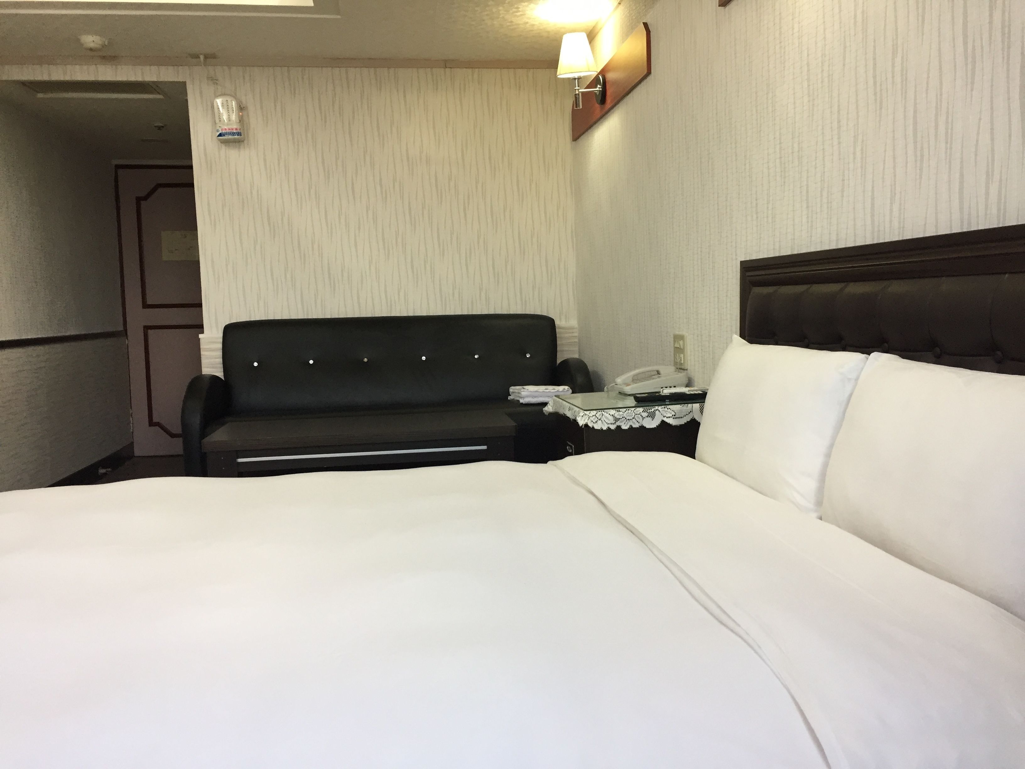 A hotel room with a bed, a small desk with a telephone, a sofa, and a wall-mounted lamp.