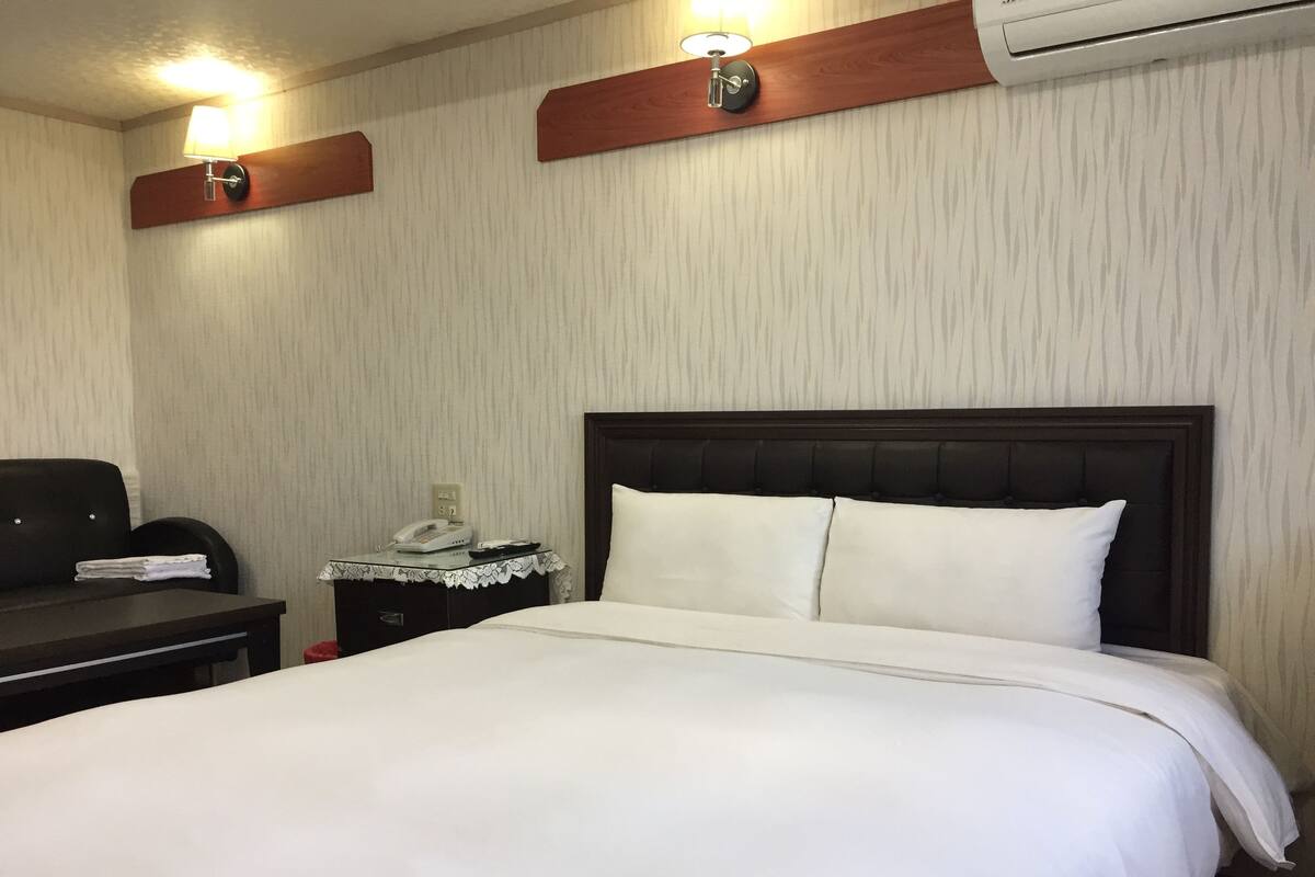 A hotel room with a large bed, a desk with a telephone, and a nightstand with a lamp.