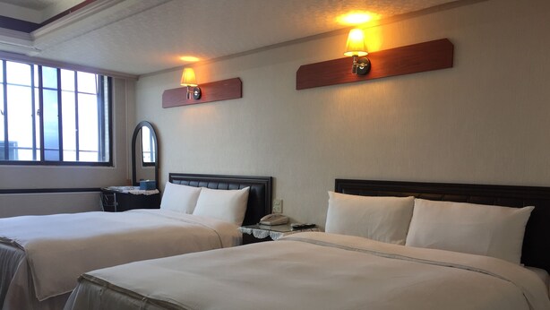 Two beds with white linens, a nightstand with a lamp, and a window with blinds. - King Lo Tung Hotel (Luodong)