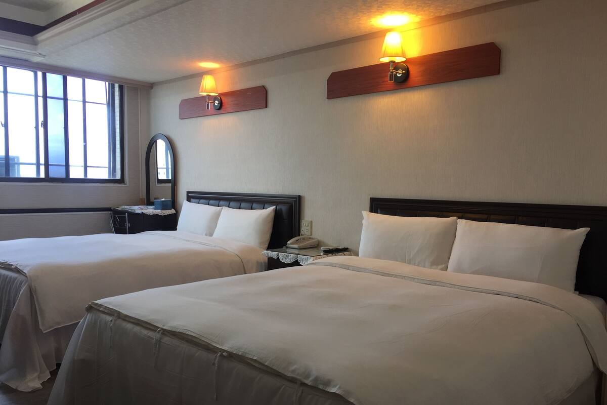 Two beds with white linens, a nightstand with a lamp, and a window with blinds.