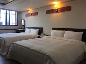 Dos camas con sábanas blancas, una mesita de noche con lámpara y una ventana con persianas.