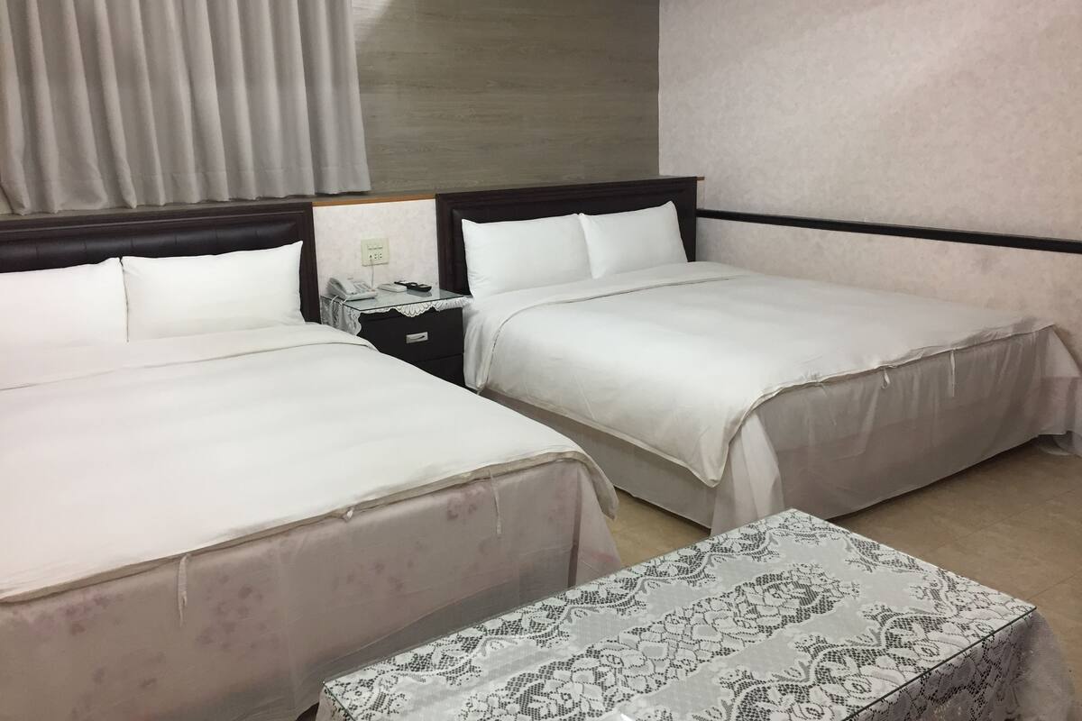 A hotel room with two beds, a bedside table with a phone and a lamp, and a small table with a patterned tablecloth.