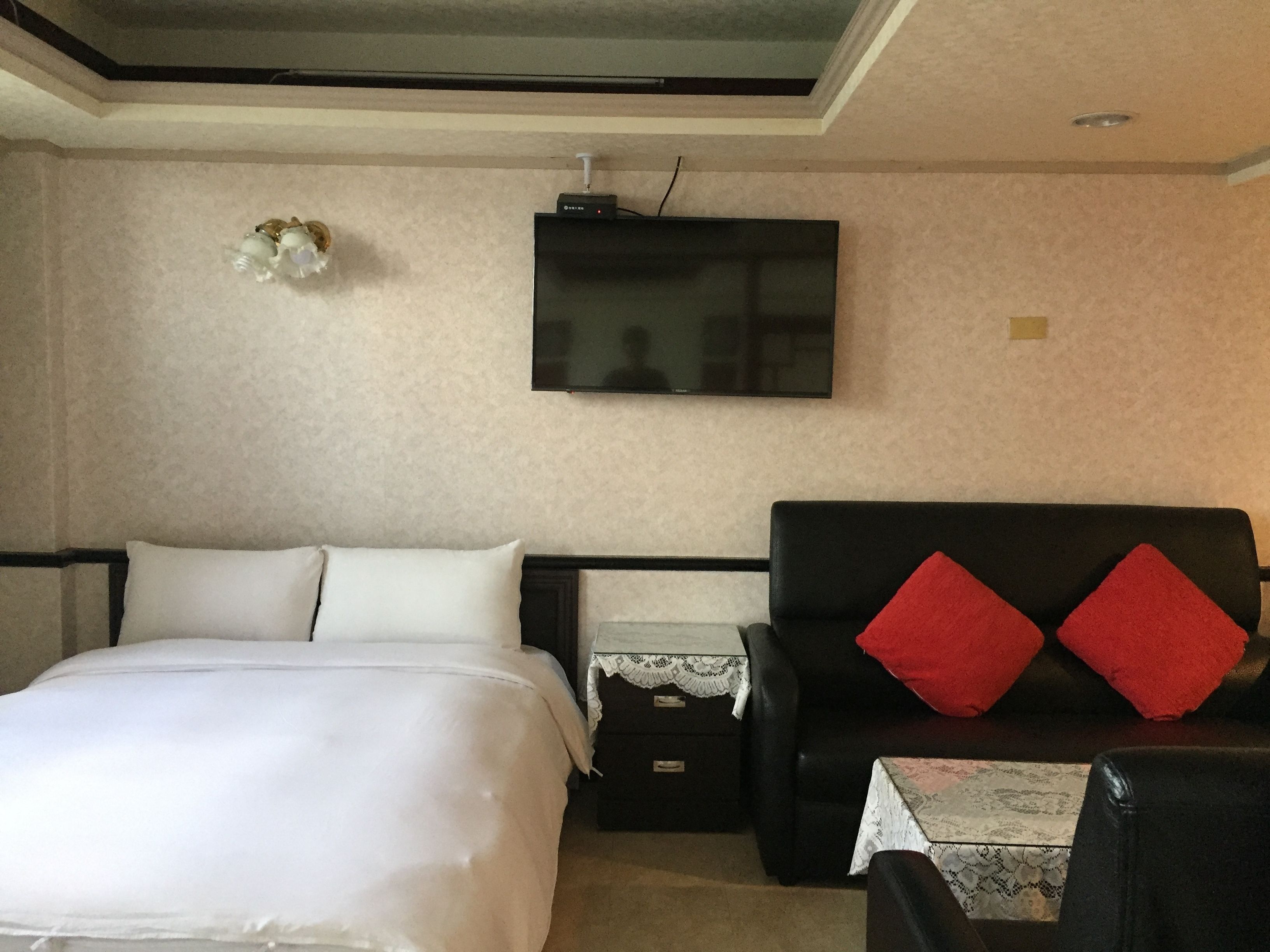 A hotel room with a bed, a sofa, a TV mounted on the wall, and a small table with a lace tablecloth.