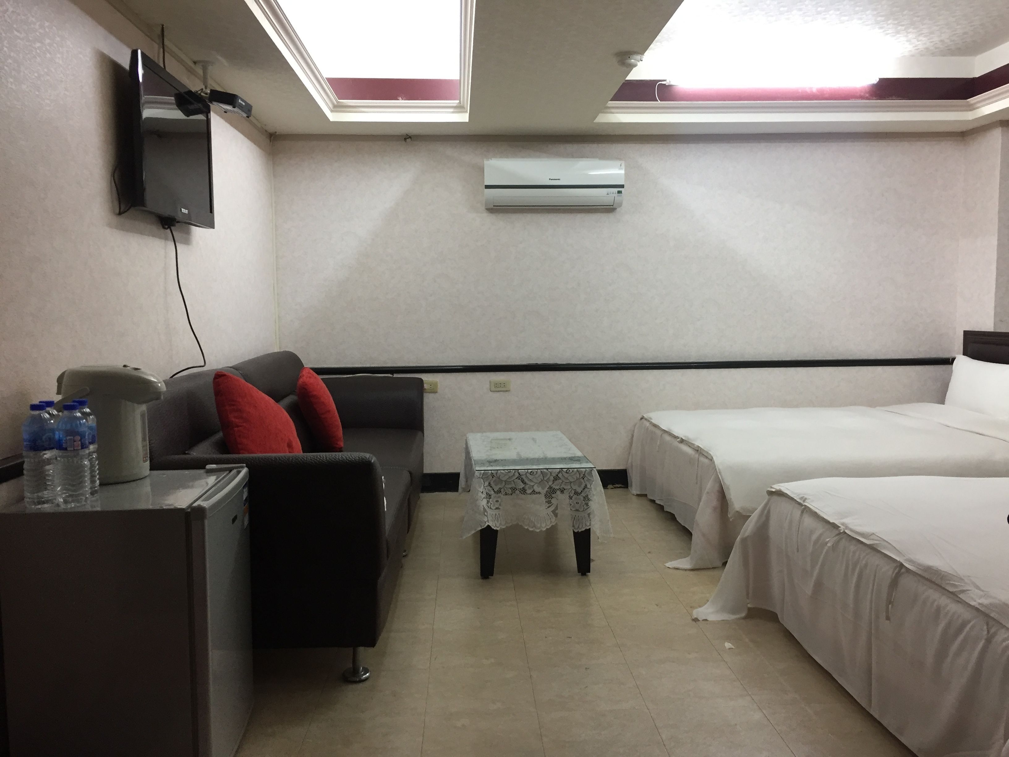 A hotel room with a bed, a desk, a chair, and a small table. There is a ceiling fan, an air conditioner, and a TV mounted on the wall.