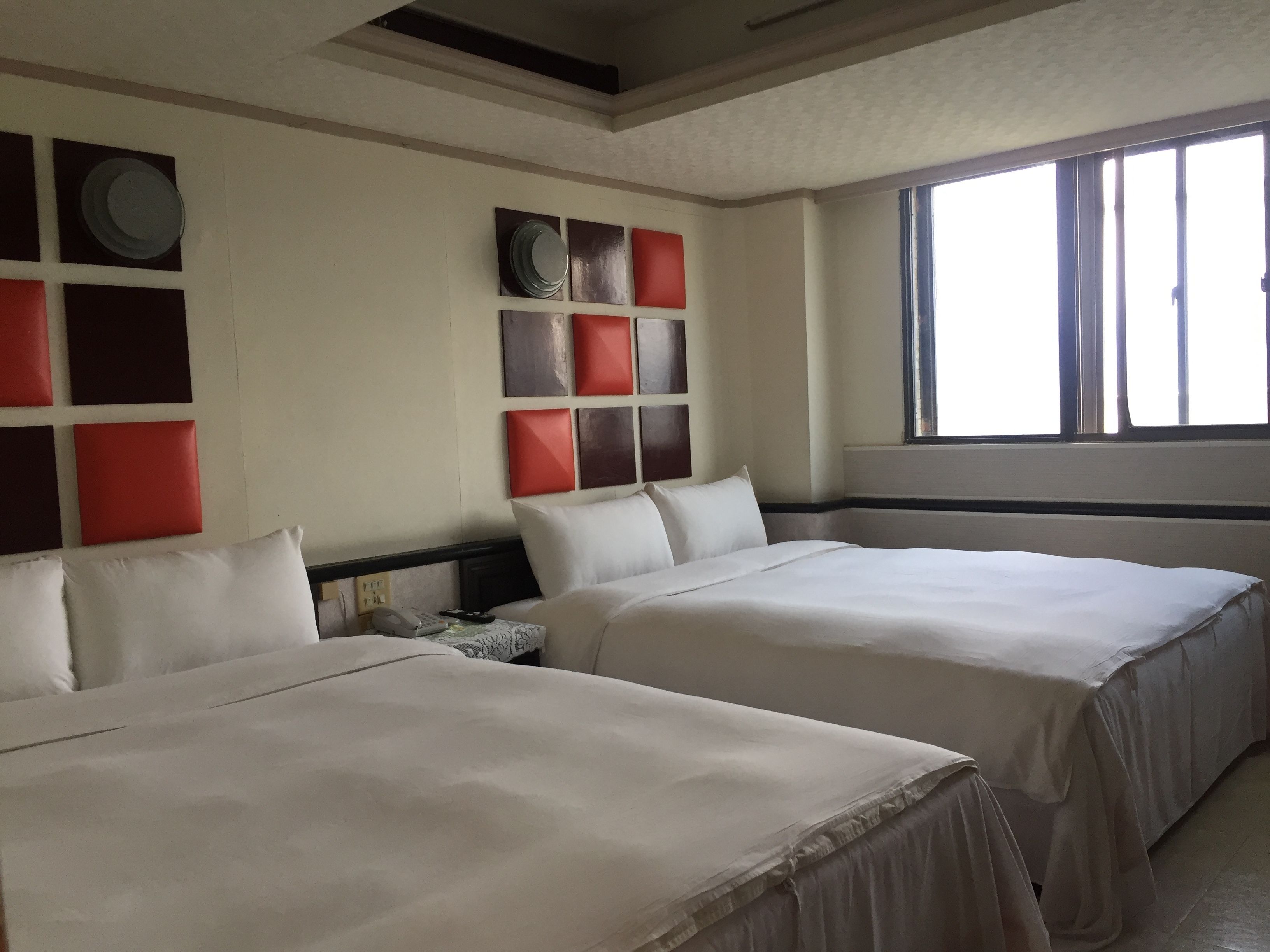 A hotel room with two beds, a large window, and a wall decorated with square panels in red and brown.