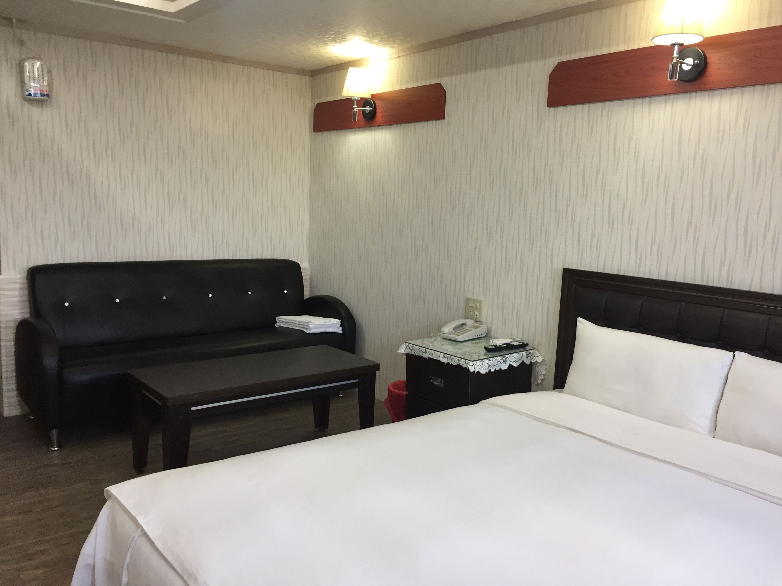 Una habitació d'hotel amb un sofà negre, una taula petita, una taula de nit amb una làmpada i un llit amb roba de llit blanca.