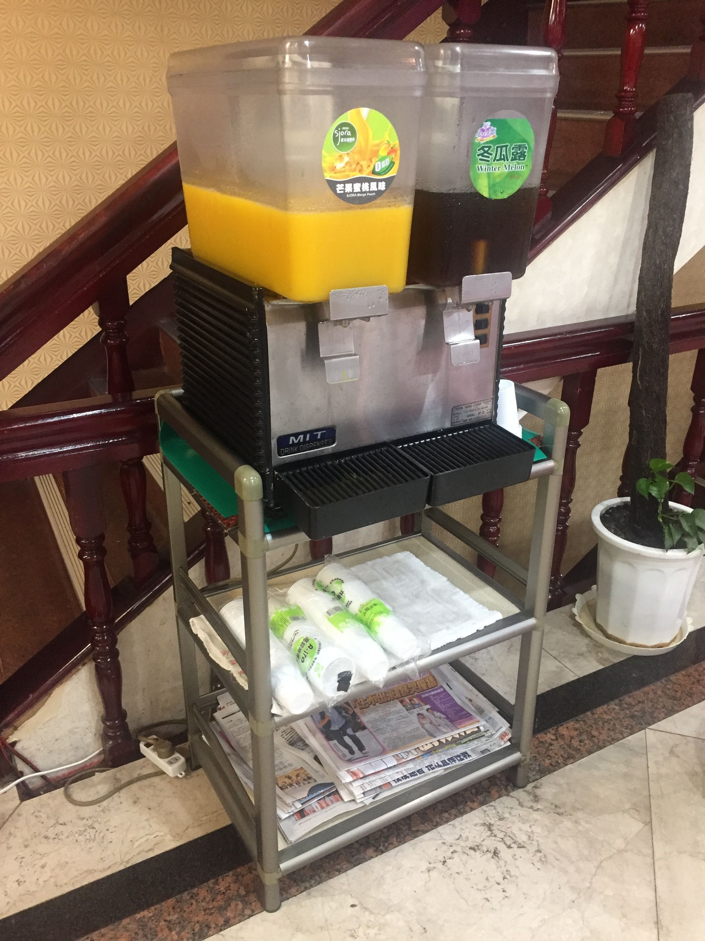 A beverage dispenser with two containers, a metal shelf with newspapers, and a potted plant.