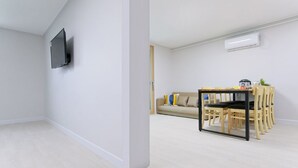 Una sala de estar moderna con un televisor de pantalla plana montado en la pared, un comedor con mesa y sillas, y un sofá color beige con cojines.