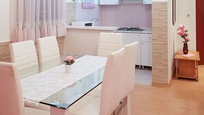 A modern kitchen with white cabinets, a gas stove, and a sink. There is a dining area with a glass table and white chairs. A small wooden table with a vase of flowers is visible in the corner. - Lovely apartment for 6 guests with A/C, WIFI, TV, terrace and pets allowed (Peroj)