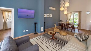 Una sala de estar moderna con un televisor de pantalla plana grande, una zona de comedor con una mesa y sillas, y un rincón de descanso acogedor con un sofá y un tapete estampado.
