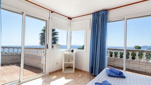 Une chambre avec une grande porte-fenêtre coulissante donnant sur un balcon, un lit avec des serviettes bleues et une petite table de chevet blanche.