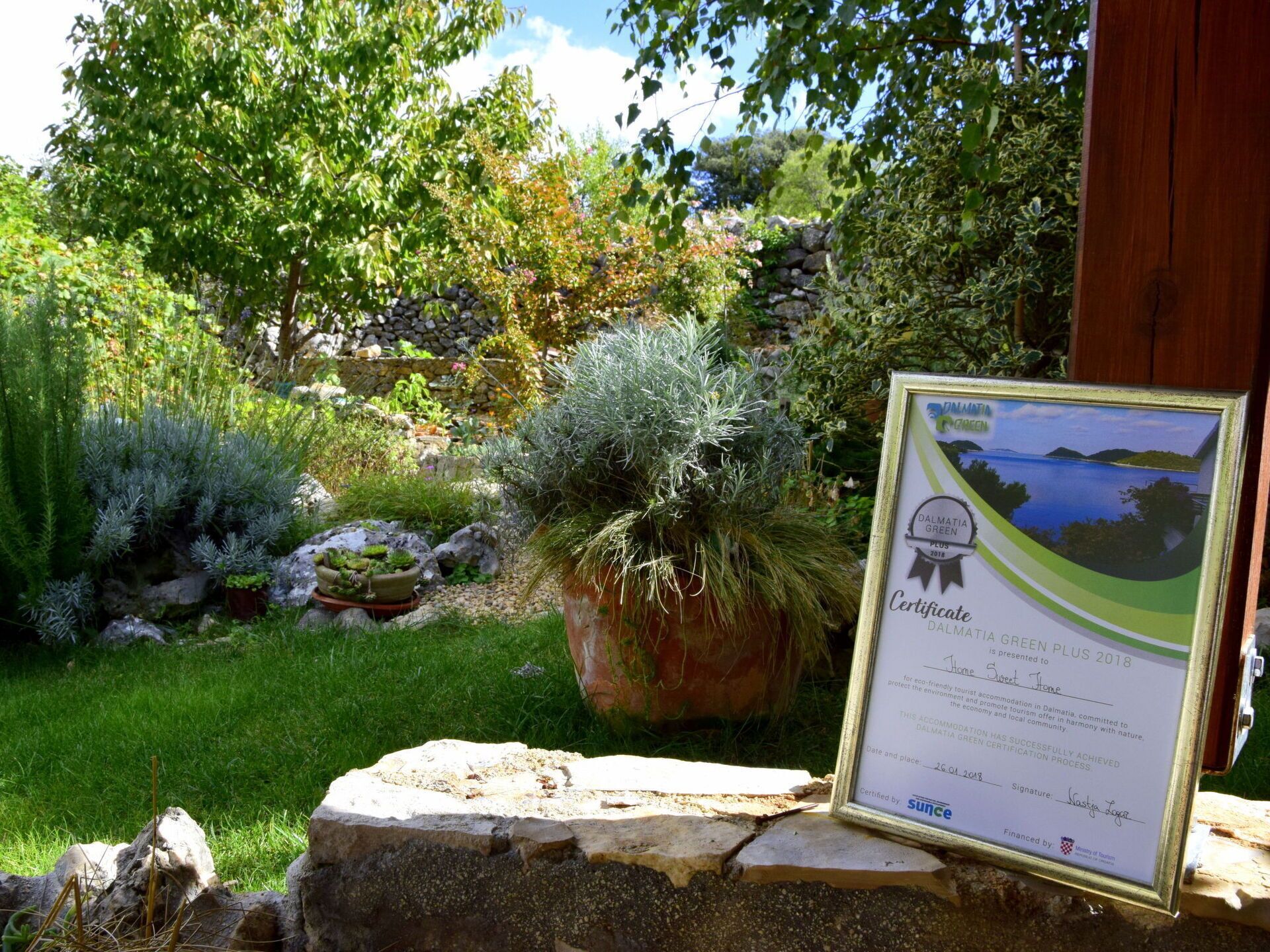 Un jardin où est affiché, encadré, un certificat attestant de l’obtention du prix « Dalmatia Green Plus 2018 », le tout placé sur une petite étagère en pierre.